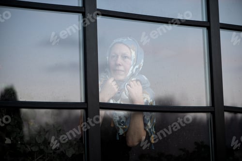 Preview: Portrait of thoughtful woman with scarf on head looking through dirty window glass