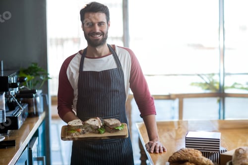 Preview: Male barista standing in cafe holding board of lettuce-topped sandwiches beside espresso machine