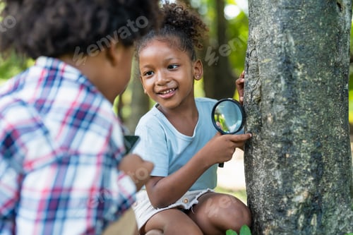 Preview: Smiling african american little girl with friends hold magnifying glass to explore on tree in park