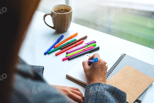 Preview: Closeup image of a woman writing on a blank notebook with colored pens and coffee cup on the table