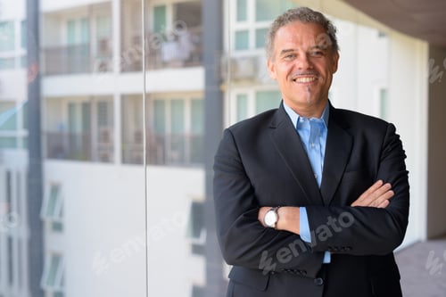 Preview: Happy mature Hispanic businessman smiling with arms crossed by the glass window of office building