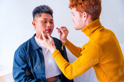 Preview: Beautiful gay couple putting on makeup painting their lips being romantic indoors at home, lgbt