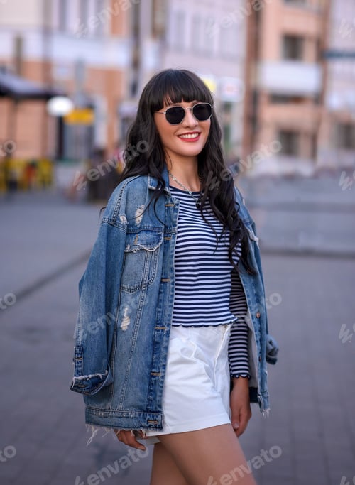 Preview: beautiful caucasian girl in a denim jacket and a short skirt and glasses on a walk in the city