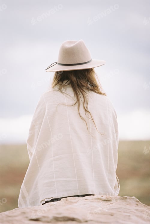 Preview: Rear view of a woman wearing a hat and wrap, sitting on a rock.