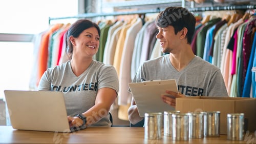 Preview: Male And Female Charity Workers At Desk With Laptop Checking Clothing Donations At Thrift Store