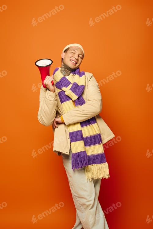 Preview: Young man with tattoos joyfully poses with a megaphone against a vibrant orange backdrop