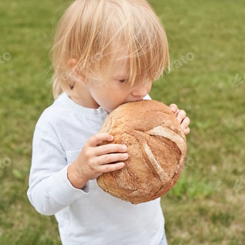 Preview: Young caucasian child enjoying large bread outdoors on grass in casual white