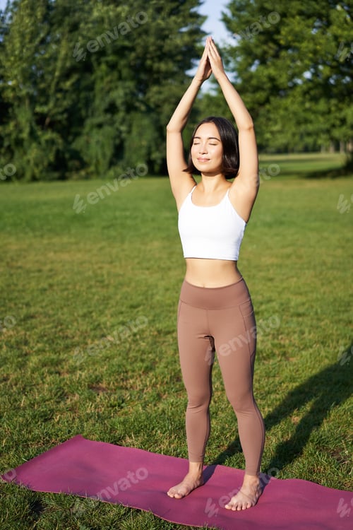 Preview: Smiling young fitness girl on rubber mat, workout in park, raising hands up in tree pose, doing