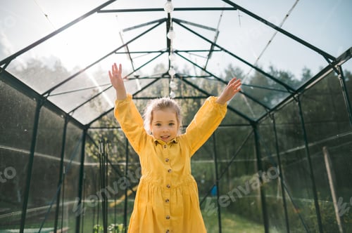 Preview: Happy little girl standing with hands raised in eco greenhouse and looking at camera.