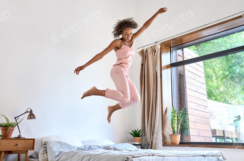 Preview: Happy carefree young African American woman wearing pajamas jumping on bed.