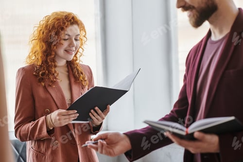 Preview: positive redhead businesswoman looking at documents near colleague in coworking office