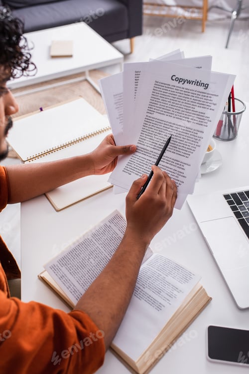 Preview: Cropped view of indian freelancer holding pencil and papers with copywriting lettering near devices