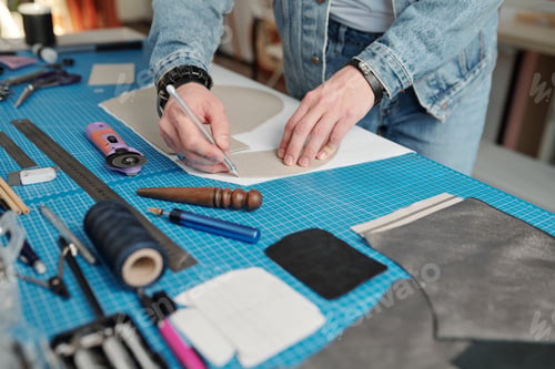 Preview: Young leatherworker in workwear outlining paper pattern over piece of suede