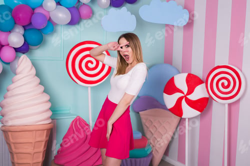 Preview: Portrait of joyful woman in pink dress on background decorated with huge candies and ice cream.