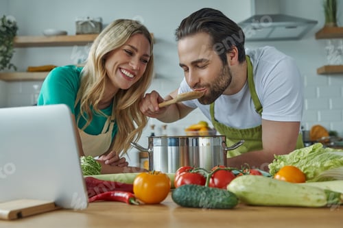 Preview: Cheerful young couple tasting something from a pan at the domestic kitchen
