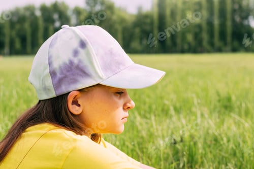 Preview: Portrait of a sad teenage girl. A preteen girl in a baseball cap sits in profile in front of a park
