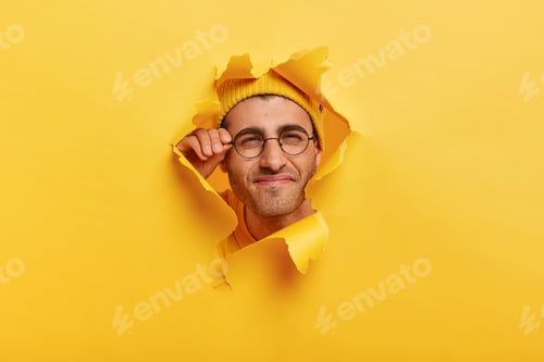 Preview: Male face in paper torn hole. Man looks curiously, has happy gaze, touches frame of glasses, poses o