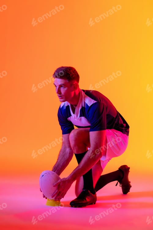 Preview: Caucasian male rugby player crouching with rugby ball over pink lighting