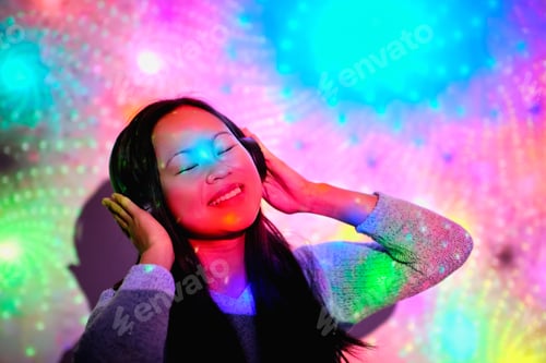 Preview: Delighted Asian woman listening to music under colorful lights
