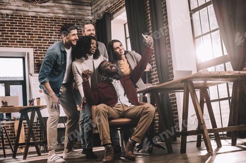 Preview: group of cheerful coworkers taking selfie using smartphone in loft office with backlit
