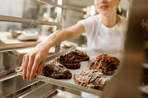 Preview: cropped shot of female baker holding tray of freshly baked croissants on baking manufacture