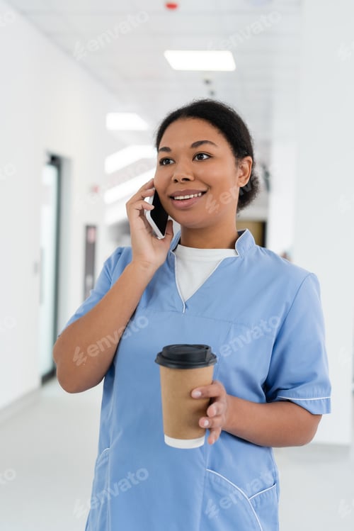 Preview: positive multiracial nurse smiling during conversation on smartphone while standing with takeaway