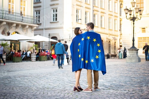 Preview: couple of young tourists, wrapped in flag of european union, standing on city square