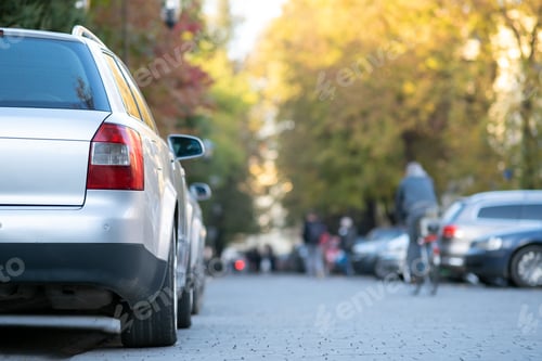 Preview: Cars parked in a row on a city street side on bright autumn day with blurred people walking on