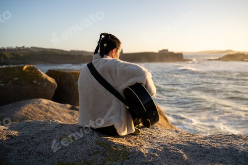 Preview: Young musician with braids sitting on rocky shoreline, embracing acoustic guitar