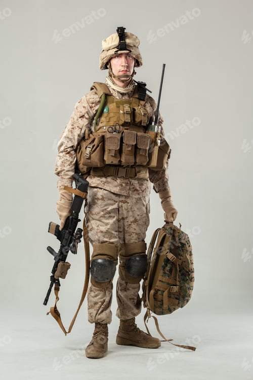 Preview: Soldier stands in uniform holding gear and weapon in a simple setting during daylight
