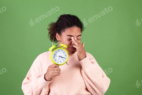Preview: Beautiful african american woman on green background with alarm clock sleepy tired exhausted yawning