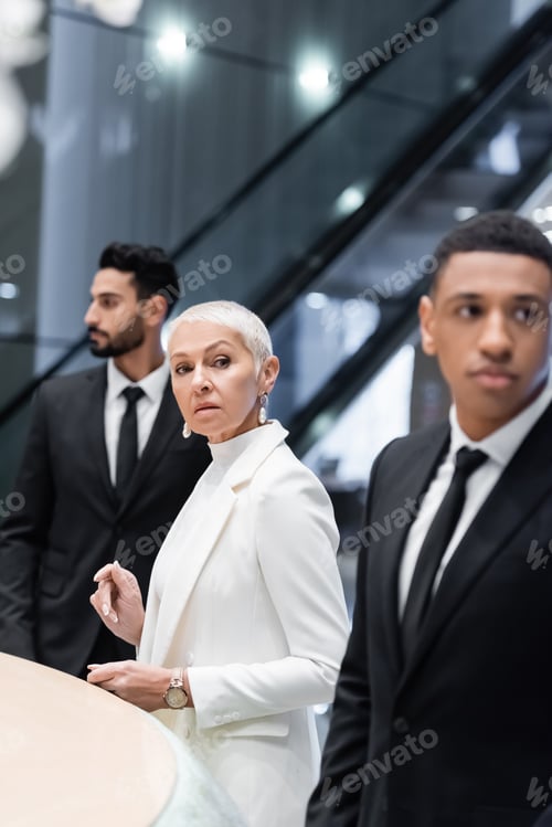 Preview: stylish senior businesswoman looking away near private interracial bodyguards in hotel lobby