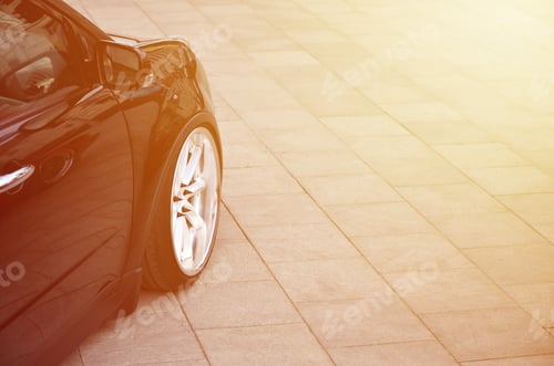 Preview: Diagonal view of a black glossy car with white wheels, which stands on a square of gray tiles