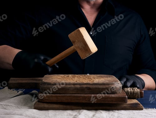 Preview: Male cook in black uniform holds a wooden hammer for beating meat on a vintage cutting board