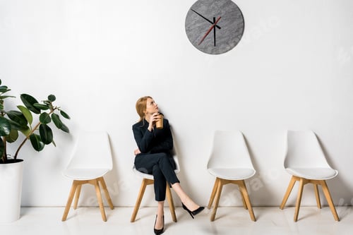 Preview: young businesswoman holding coffee to go and looking at clock while waiting in line