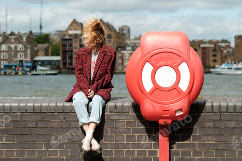 Preview: Woman sitting on brick banister beside lifebuoy in London riverside.