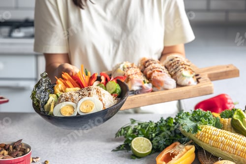 Preview: Bowl with vegetables and rice and spring rolls in female hands in the kitchen.
