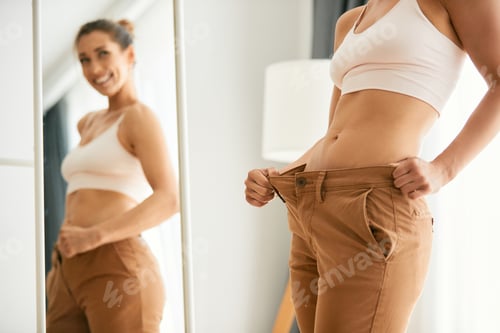 Preview: Slim woman in overweight pants looking herself in a mirror after loosing weight.