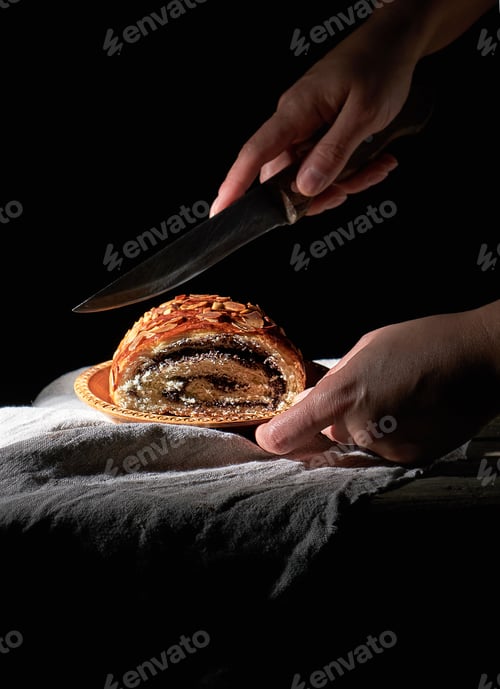 Preview: Female hands cut baked roll with poppy seeds on a wooden plate
