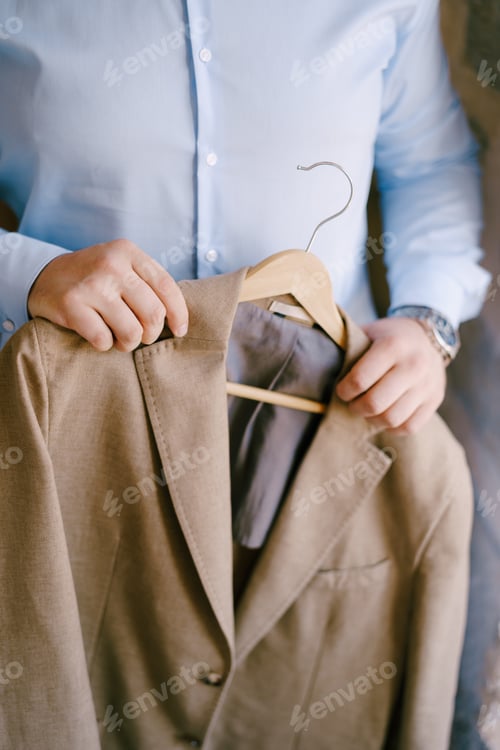 Preview: Groom in a blue shirt holds a brown jacket on a hanger