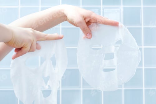 Preview: Female hands holding two moisturizing mask sheets close-up against background of blue tiles.