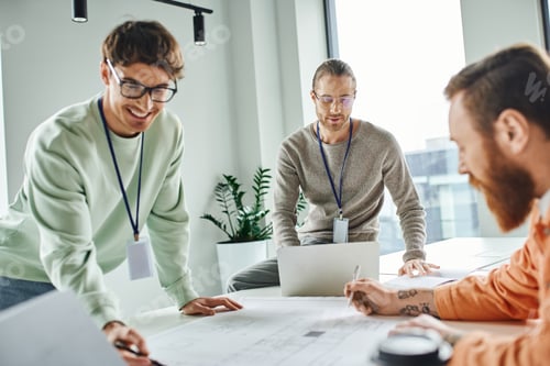 Preview: smiling architect pointing at blueprint near colleagues and laptop while working in coworking space