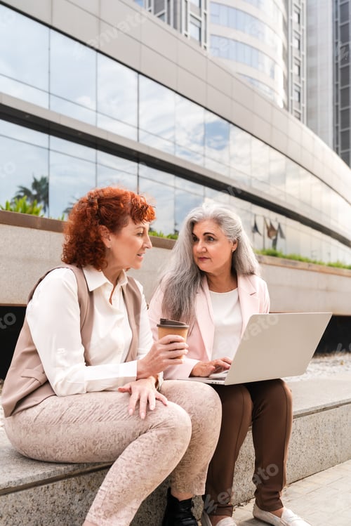 Preview: Two senior businesswomen talking while working on laptop sitting outside modern office building