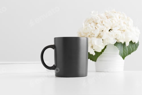 Preview: Black mug mockup with a book and a hortensia in a pot on a white table.
