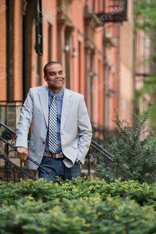 Preview: A man in a jacket and tie on a city street outside a terrace of brownstone houses.