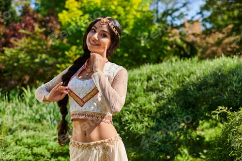 Preview: cheerful indian woman in elegant ethnic wear posing at camera in green summer park