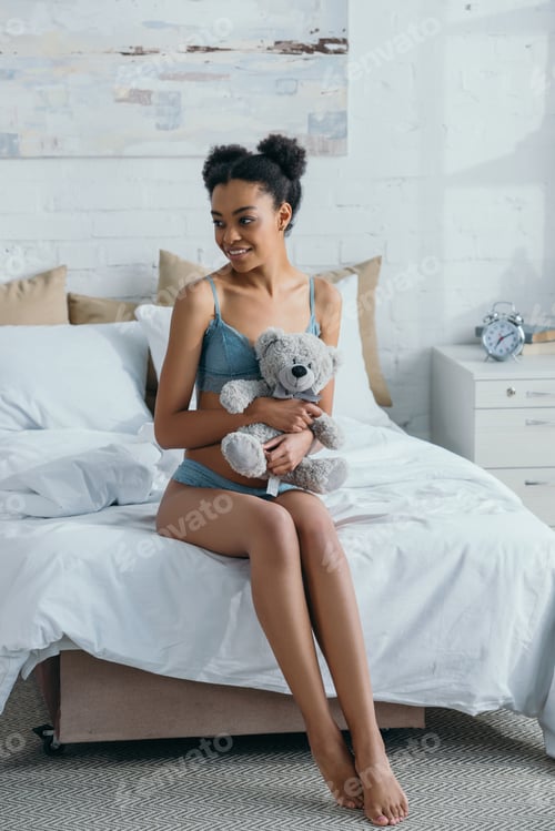 Preview: happy african american girl in lingerie holding teddy bear in bedroom in morning