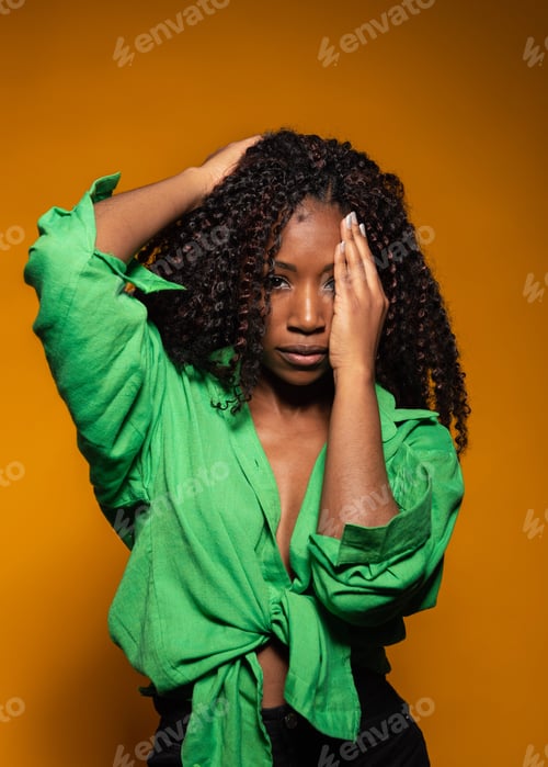 Preview: Serious Hispanic woman in green shirt covering one eye with a hand in studio on yellow background