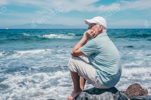 Preview: Carefree senior bearded man with cap sitting barefoot on the rocky beach enjoying summer