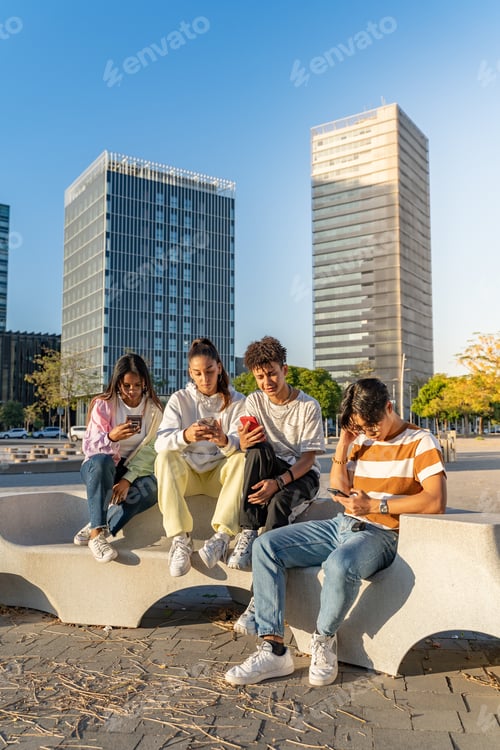 Preview: Group of happy teenage friends looking the phones in a bench in the city street.